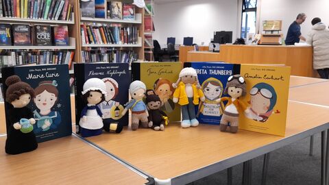 Five crocheted characters stand next to books about the characters on a table in a library