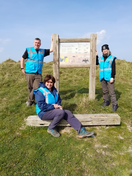 3 people standing around a wooden noticeboard on a sand dune