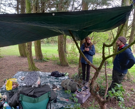 2 people standing under a tarp in the rain surrounded by trees